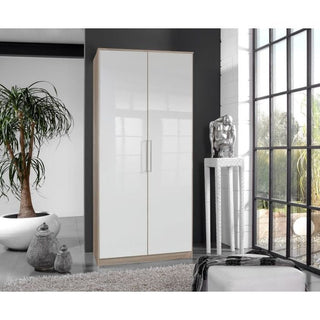 A modern room with the GEMA 2 Door Wardrobe in Oak and White Gloss (90cm), a tall potted plant, floor vases, a small table with a silver sculpture, and large windows filling the space with natural light.