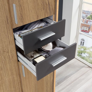 Two open drawers in the FENTON 4 Door 4 Drawer Mirrored Wardrobe, finished in oak and graphite, reveal neatly folded clothes and personal items. The wardrobe stands by a window with a view of buildings outside.