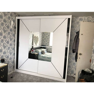 The BARTHOLOMEW 2 Door Mirrored Sliding Wardrobe in white (120cm) stands against floral wallpaper, reflecting the bed and decor. A coat hangs on an open door, with a small dresser visible to the left.
