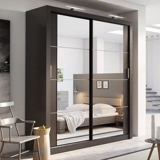 A modern bedroom showcases the CHOICEWARDROBE Allen Wardrobe, a full-height matt black freestanding unit with two sliding mirrored doors reflecting a neatly made bed, wall art, and minimalist decor. A chair and small table sit in the foreground.