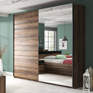 The Biloxi Mirrored Wardrobe 2 Sliding Doors 200cm in Monastery Oak, with one mirrored and one solid door, stands against a green bedroom wall and reflects a bed, window, and modern light fixture.