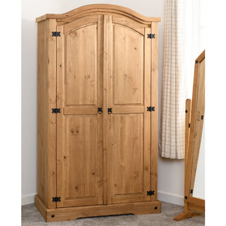 The Central Wooden Wardrobe With 2 Doors In Oak, featuring decorative black hinges, a curved top, and natural wood finish, stands against a white wall near a tall mirror and a window with white curtains.