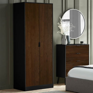 A bedroom featuring the CHOICEWARDROBE Akron 2 Door Wooden Wardrobe in walnut and black, a matching three-drawer dresser with a round mirror above, white branch vases, and part of a bed with a grey headboard.