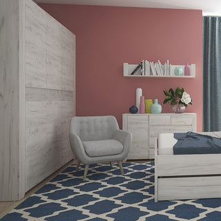 A bedroom with a light wood bed, dresser, CHOICEWARDROBE Alink Large 3 Door Wardrobe in white oak effect, gray armchair, navy and white geometric rug, pink accent wall, shelf with books and decor, and blue curtains.