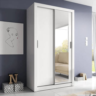 The CHOICEWARDROBE Aliso 120cm White Sliding Door Wardrobe with Mirror stands against a blue wall, featuring one mirrored and one plain door. A line-drawing portrait, boxes, and stacked magazines are nearby, with the mirror reflecting a window and beige sofa.