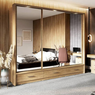 A modern bedroom with wood-paneled walls, a CHOICEWARDROBE Aliso 250cm 3 Door Sliding Mirrored Wardrobe in Shetland Oak with LED lighting and storage drawers, a bed with white bedding, a desk and chair, and plants in vases on the carpet.