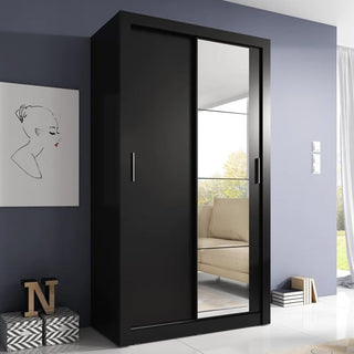 A CHOICEWARDROBE Aliso Sliding Door Wardrobe – 120cm in Matt Black, with two smooth sliding doors (one mirrored) and adjustable storage shelves, stands against a gray wall beside minimalist décor, stacked books, and a beige chair’s reflection.