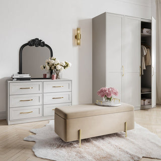 A modern bedroom features a white dresser, a black-framed mirror, a vase of white flowers, the CHOICEWARDROBE Allen 1 Door Slim Wooden Wardrobe in white with gold handle, a beige bench with gold legs, and a light cowhide rug on wood floors.