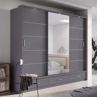The CHOICEWARDROBE Allen 250cm 3 Door Sliding Mirrored Wardrobe in matt grey, featuring drawers, shelves, and hanging rails, stands against the wall; its mirrored doors reflect a bedside table and bed, with a chair draped in plaid nearby.