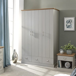 A modern bedroom with the CHOICEWARDROBE Alverton 3 Door Wardrobe in grey with an oak effect top and drawers, complemented by a matching nightstand, blue curtains, a framed leaf print, and a glass vase.