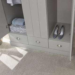The CHOICEWARDROBE Alverton 3 Door Wardrobe in grey with oak effect top and drawers displays folded clothes, a towel, and grey shoes on shelves above three closed drawers, standing on a light-colored carpeted floor.