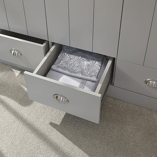A partially open drawer in the CHOICEWARDROBE Alverton 3 Door Wardrobe – Grey with Oak Effect Top & Drawers reveals neatly folded, embroidered bedding; other drawers are slightly open, and the room features a carpeted floor.