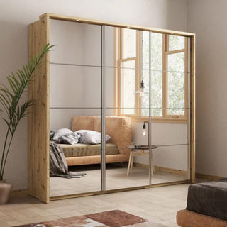 The CHOICEWARDROBE Amarillo 240cm Natural Oak Sliding Wardrobe with 3 full mirror doors and a Scandinavian wood finish stands against a wall, reflecting a beige sofa, side table, hanging light, window, and nearby green plant in a modern room.