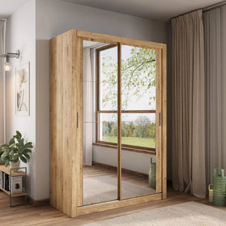 The CHOICEWARDROBE Ames Mirrored Sliding Door Wardrobe in Oak Shetland finish (150cm, 2 doors) stands in a bright, carpeted room by a window, with a plant, green basket nearby, and a framed picture on the wall.