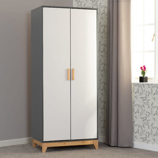The CHOICEWARDROBE Arthur 2 Door Wardrobe—white and grey with oak legs in Scandinavian design—is placed by a window with gray curtains and patterned wallpaper in a carpeted room, with a small pink flower on the windowsill.