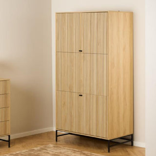 The CHOICEWARDROBE Attica 2 Door Wooden Wardrobe (99cm, Oak & Matt Black finish) features a fluted design and metal frame base, providing stylish bedroom storage against a beige wall and parquet floor.