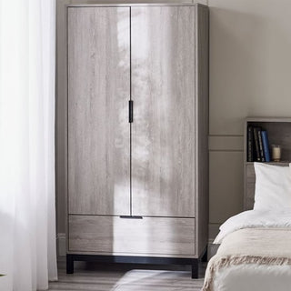 The CHOICEWARDROBE Baara 2 Door Wardrobe in light grey ash effect with black metal legs and drawer stands by a bed and nightstand in a sunlit bedroom, offering stylish modern industrial storage. Light filters through a sheer curtain on the left.