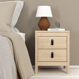 A bedside table from the CHOICEWARDROBE Bellevue 3 Piece Bedroom Furniture Set in oak effect stands by a bed with beige bedding, holding a brown and white lamp, a closed "SCULPTURE" book, and a clear glass of water.