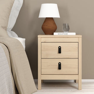 A CHOICEWARDROBE Bellevue bedside table with oak effect finish from the 3 Piece Bedroom Furniture Set stands by the bed, topped with a brown and white lamp, the book SCULPTURE, and a glass of water against a beige wall.