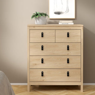 A CHOICEWARDROBE Bellevue 3 Piece Bedroom Furniture Set in oak effect stands against a beige wall, featuring a 5 drawer chest with black handles. On top are a potted plant, two books, and a framed abstract line drawing hanging above.