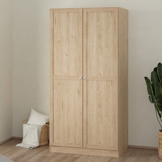 The CHOICEWARDROBE Bensalem 2 Door Wooden Wardrobe in oak finish stands against a light wall, with a plant and a basket of pillows and a blanket beside it, offering classic design and spacious hanging storage for your bedroom.