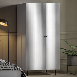 A CHOICEWARDROBE Bienne White Fluted Wooden Wardrobe with 2 doors, drawers, and black metal legs stands beside a green potted plant, with a floor lamp and part of a bed with a patterned blanket visible in the foreground.