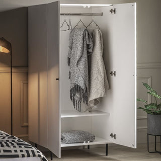 The CHOICEWARDROBE Bienne White Fluted Wooden Wardrobe stands open, displaying three gray garments and a folded item inside. Beside it are a green potted plant, a standing lamp, and part of a bed.