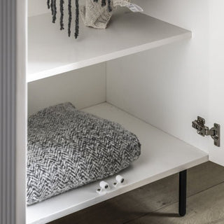 The CHOICEWARDROBE Bienne White Fluted Wooden Wardrobe stands open, showing shelves with a folded grey herringbone blanket below and a plush toy above; white hinge and black metal legs add modern style.