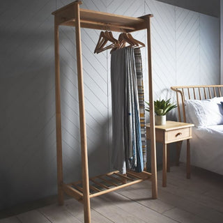 The CHOICEWARDROBE Burbank Open Wardrobe, a solid oak Scandinavian freestanding garment rack with shelf storage, stands next to a bed and small wooden nightstand with a potted plant in a minimally decorated bedroom.