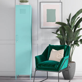 A modern interior featuring the CHOICEWARDROBE Caches Metal Locker Wardrobe in spearmint, a dark green velvet armchair with a beige cushion, a large leafy plant, and framed abstract art on a white-paneled wall.