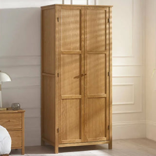 The CHOICEWARDROBE Calliope 80cm 2 Door Solid Wooden Wardrobe in a natural oak finish, featuring a hanging rail and shelf, stands against a white wall in a bright bedroom, with sunlight casting shadows and a bedside table and lamp nearby.