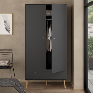 The CHOICEWARDROBE Casey Wooden Wardrobe—2 doors, 2 drawers, dark grey with oak legs and brass handles—stands against a beige wall. One door is partly open, showing hanging clothes and a bag. Nearby are a chair, wall art, and window.