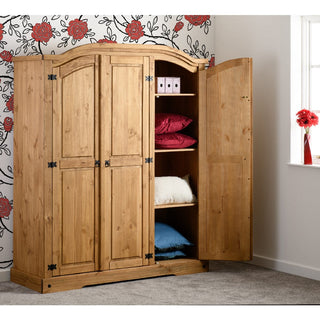 The CHOICEWARDROBE Central 3 Door Wooden Wardrobe in solid pine features shelves holding folded blankets, pillows, and storage boxes. Its rustic farmhouse design stands on a carpeted floor against floral wallpaper near a window.