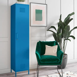 The CHOICEWARDROBE Cerritos 38cm Metal Wardrobe in bold blue stands beside a green velvet chair with a beige cushion, set against paneled white walls, framed artwork, and a large plant.