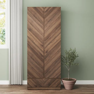 The CHOICEWARDROBE Cianna Royal Walnut 2 Door Wardrobe with Drawer, featuring a chevron pattern luxury finish, stands against a pale green wall with a potted plant on the right and a white curtain by the window on the left.
