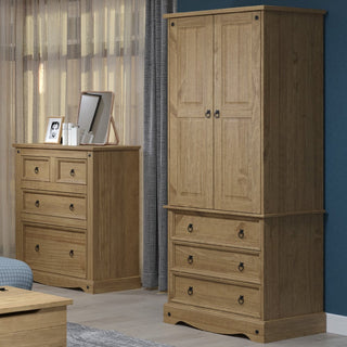 The CHOICEWARDROBE Consett 2 Door 3 Drawer Solid Pine Wardrobe with black metal handles stands on the right in a blue-walled bedroom with sheer curtains. A chest of drawers on the left displays photos, a mirror, and small decor items.