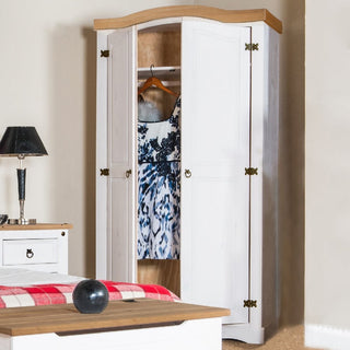 The CHOICEWARDROBE Consett 2 Door Wooden Wardrobe (100cm, White & Oak finish, classic curved top, rustic metal hinges) stands in a bedroom with its doors partly open to show a dress inside, near a bed, nightstand, lamp, and décor.