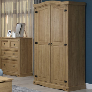 A CHOICEWARDROBE Consett 2 Door Wooden Wardrobe in natural pine with a rustic curved top and metal hinges stands by a matching dresser, mirror, and photo frame against a blue wall with sheer curtains.