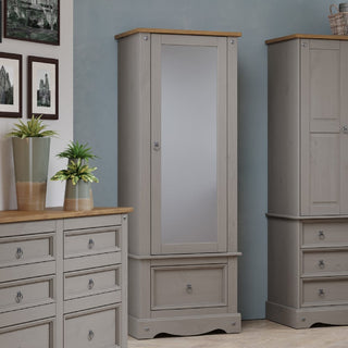 The CHOICEWARDROBE Consett 60cm Grey & Oak Wooden 1 Door Mirrored Wardrobe with Drawer stands between a plant-topped dresser and a cabinet against a light blue, picture-adorned wall, offering compact, stylish bedroom storage.