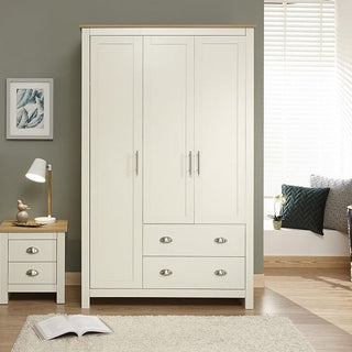 The CHOICEWARDROBE Crick Cream & Oak 3 Door Wardrobe with shaker style and deep drawers stands beside a matching bedside table. The room has a grey wall, rug, and blinds on the window that let in natural light.
