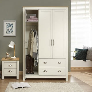 The CHOICEWARDROBE Crick Cream & Oak 3 Door Wardrobe with 2 deep drawers stands open, showing clothes, boots, and towels. Nearby is a white nightstand with a lamp, book, and clock. A window and cushioned seat are in the background.