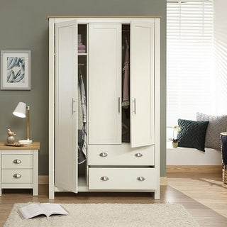 The CHOICEWARDROBE Crick Cream & Oak 3 Door Shaker Wardrobe with 2 deep drawers stands open, showing hanging clothes inside. It's beside a matching nightstand and lamp, against a gray wall in a bright bedroom with a window seat and pillows.