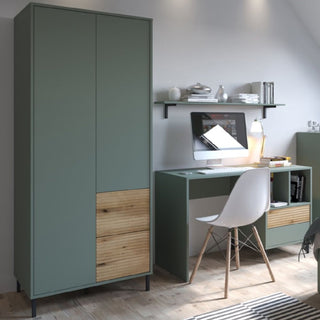 A minimalist home office features the CHOICEWARDROBE Danville 2 Door Wooden Wardrobe in Sage Green & Oak, paired with a matching desk holding essentials. A shelf above displays books and decor, and a white chair sits in front for seating.