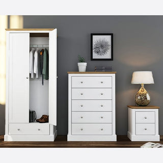 The CHOICEWARDROBE Devan 2 Door Wardrobe, 80cm wide in white with an oak top and drawer, stands open by a chest of drawers and bedside table against a gray wall, displaying hanging clothes and shoes. A lamp and framed artwork rest atop the furniture.