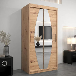 The CHOICEWARDROBE Eden 100cm Oak Mirrored Wardrobe, featuring unique curved mirror panels and silver handles, stands against a white wall beside a black nightstand with dried flowers. A TV and lamp are visible in the background.