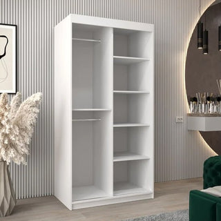 The CHOICEWARDROBE Eden 100cm White Mirrored Sliding Door Wardrobe, featuring modern curved mirrors, shelves, and a hanging rail, stands in a stylish room with light wood floors, a green velvet sofa, decorative grass, and a circular wall mirror.