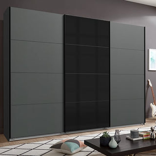 The CHOICEWARDROBE Ernesto 3 Door Sliding Wardrobe, featuring a matt graphite and black glass finish, stands against a grey wall. A guitar, framed art, and a decorated coffee table enhance the contemporary bedroom setting.