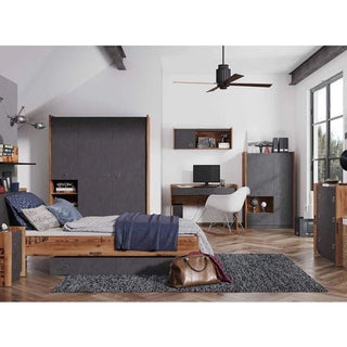 A modern bedroom with a wooden bed, gray bedding, desk and computer, white chair, bookshelves, and a CHOICEWARDROBE Fairbourne 3 Door Wardrobe in raw steel & reclaimed wood effect. A brown leather bag sits on a gray rug by the large window.