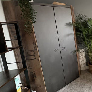 The CHOICEWARDROBE Fairbourne L-Shaped Corner Wardrobe in raw steel and reclaimed wood effect stands on a carpeted floor, framed by leafy plants and a shelving unit filled with books and decor items.
