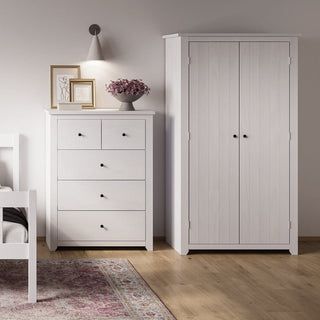 A bedroom corner with a white dresser, bed frame, wall lamp, framed art, flowers in a vase, patterned rug on wooden floor, and the CHOICEWARDROBE Havanan 2 Door White Wooden Wardrobe featuring a modern vertical panel design.